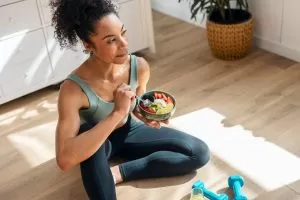 An athletic woman is shown eating a healthy fruit bowl while sitting on the floor in her kitchen at home.