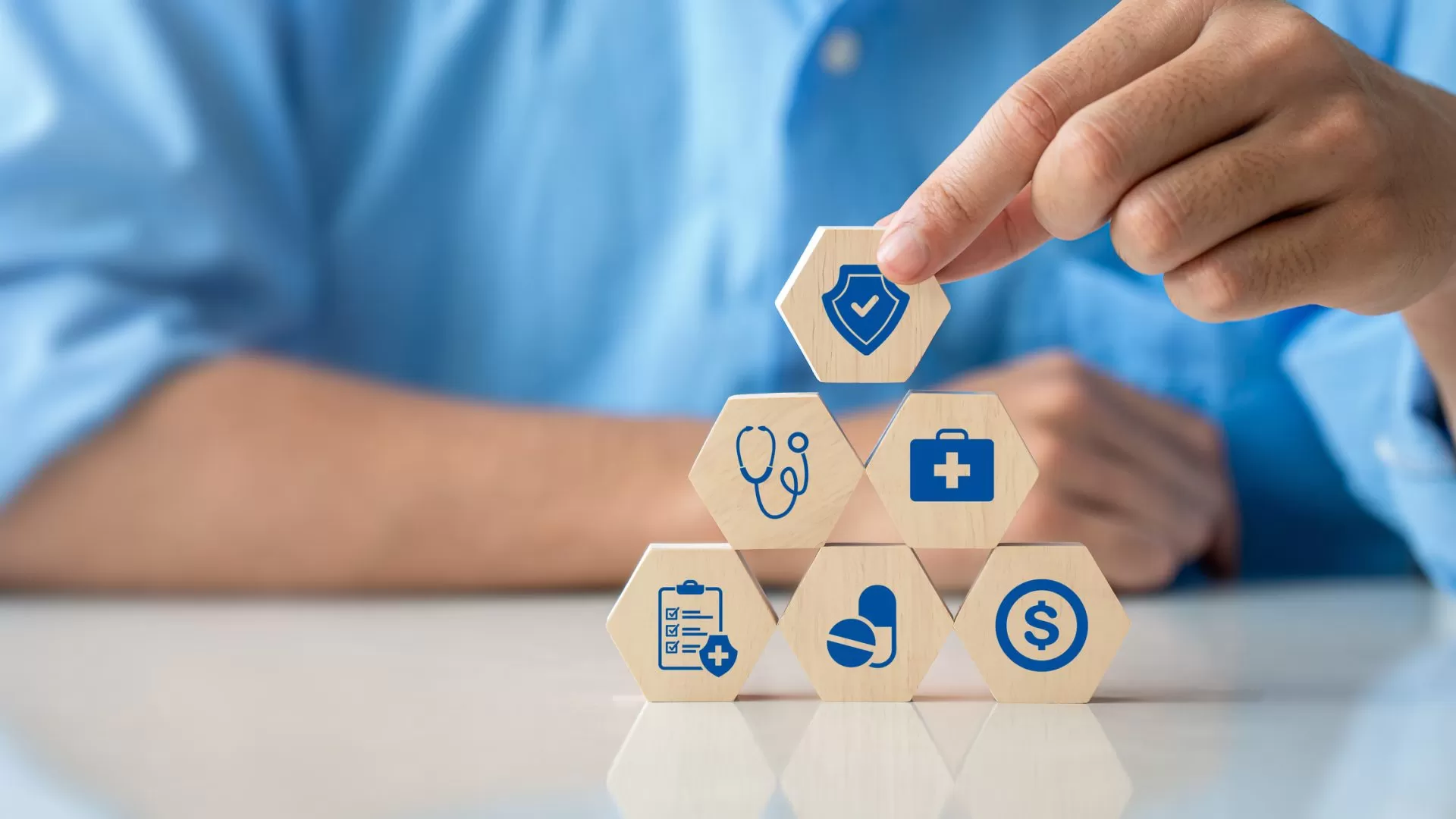 Close-up of a hand holding wooden cube with a healthcare medical icon on it, being stacked on top of five other wooden cubes with healthcare symbols.